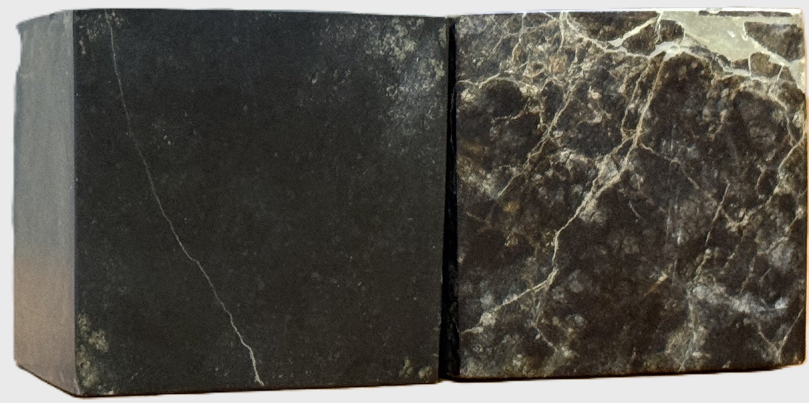 Two saw-cut cubes of ophiolite rock: fresh and near-black on the left, browned and cut by white carbonate veins after CO₂ mineralisation on the right